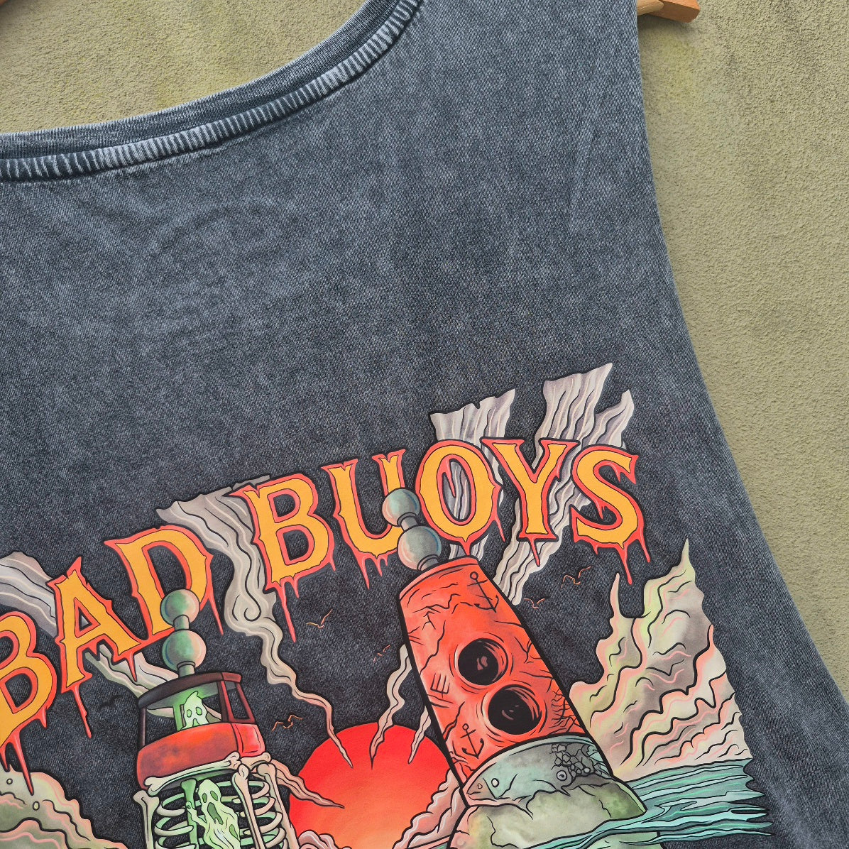 Close-up of a dark black acid-wash tank top featuring a colorful nautical print with the text 'BAD BUOYS' and illustrations of a skulls and marker buoys