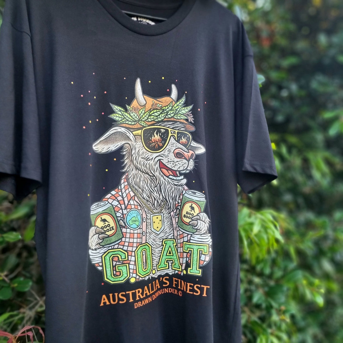 Black t-shirt featuring G.O.A.T. - Australia's Finest graphic by Drawn Downunder. Funny Aussie goat wearing a cork hat, sunglasses with campfire reflection, flannel shirt and holding two Roo Booze beer cans. Hand-drawn Australian art tee hanging outdoors.